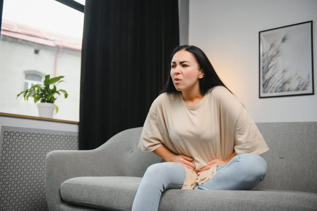 Femme assise sur un canapé, les mains sur le bas-ventre, le visage crispé par la douleur